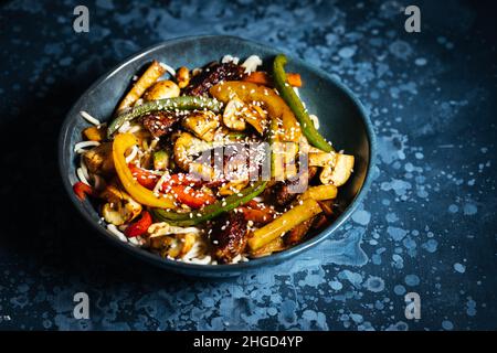 Wok avec nouilles, poivrons, champignons, viande de dinde et différents légumes et graines de sésame dans un bol bleu sur fond sombre Banque D'Images