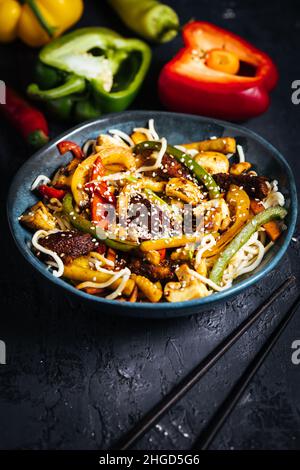 Wok avec nouilles, poivrons, champignons, viande de dinde et différents légumes et graines de sésame dans un bol bleu sur fond sombre Banque D'Images