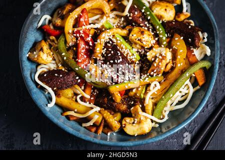 Wok avec nouilles, poivrons, champignons, viande de dinde et différents légumes et graines de sésame dans un bol bleu sur fond sombre Banque D'Images