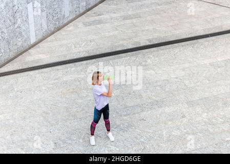 Par dessus la jeune femme sportive en forme d'activewear boire de l'eau rafraîchissante de la bouteille tout en se reposant après l'entraînement en plein air sur la place urbaine Banque D'Images