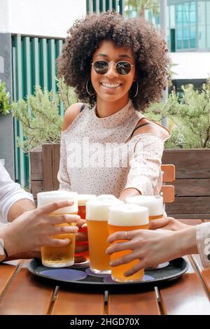 Gaie femme noire avec des cheveux bouclés dans des vêtements élégants et des lunettes de soleil assis à la table et se clinquant avec un verre de bière avec des amis tout en passant Banque D'Images