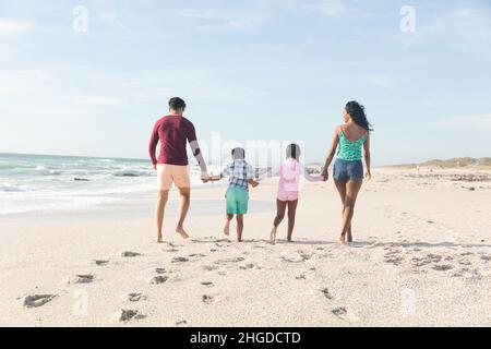 Toute la famille multiraciale tient les mains ensemble marchant sur le sable avec des empreintes de pas à la plage Banque D'Images