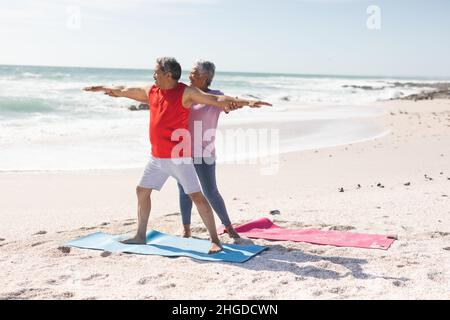 Femme biraciale senior aidant l'homme avec guerrier pose posture de yoga pratiquant à la plage contre le ciel Banque D'Images