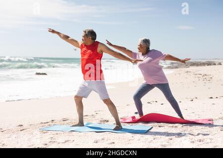 Biracial couple senior pratiquant le guerrier pose le yoga sur des tapis à la plage contre le ciel pendant la journée ensoleillée Banque D'Images