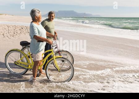 Pleine longueur de couple multiracial senior avec des vélos debout sur la plage pendant la journée ensoleillée Banque D'Images