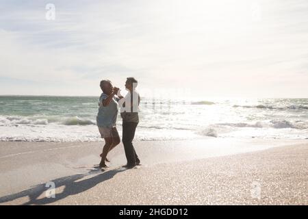 Vue latérale sur le couple multiracial senior heureux dansant romantique à la plage par beau temps Banque D'Images