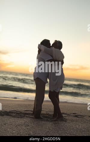 Vue latérale sur une longue distance d'un couple biracial senior heureux s'embrassant les uns les autres tout en se tenant à la plage Banque D'Images