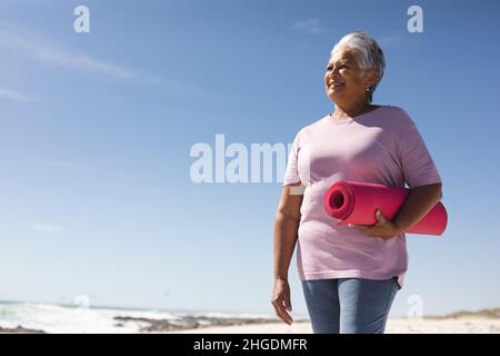 Femme biraciale senior souriante tenant un tapis de yoga rose tout en regardant la plage contre le ciel bleu Banque D'Images