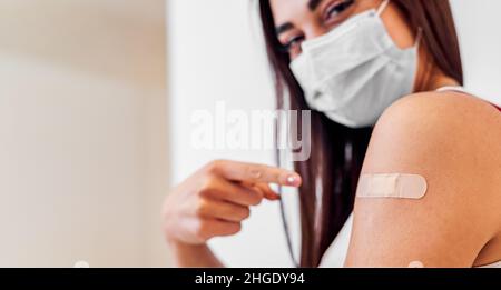 Jeune femme montrant avec le doigt le bandage sur son épaule de bras portant un masque de protection fait avec la vaccination.La femme vient de se faire vacciner Banque D'Images