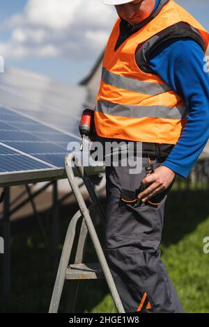 Un ingénieur qui installe et entretient des panneaux solaires se tient sur un escabeau et sort une clé de sa poche Banque D'Images