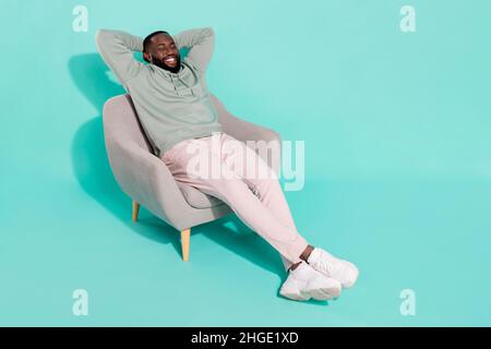 Vue sur toute la longueur du corps d'un sympathique gars de rêve assis dans un fauteuil reposant isolé sur un fond bleu turquoise clair de couleur turquoise Banque D'Images