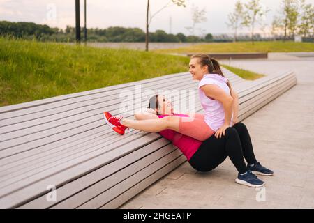 Gros plan de l'entraînement de la jeune femme gras avec instructeur de fitness gai en utilisant le banc à la salle de gym en plein air dans le parc de la ville. Banque D'Images