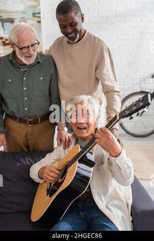 Homme asiatique senior jouant de la guitare acoustique et chantant près d'amis multiethniques à la maison Banque D'Images