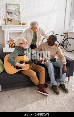 Homme asiatique chantant près d'amis multiethniques avec de la guitare acoustique sur le canapé à la maison Banque D'Images