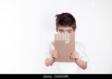 Un petit garçon couvre la moitié de son visage avec un livre et montre seulement ses yeux sur fond blanc. Banque D'Images