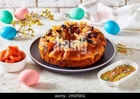 couronne de pain de pâques parsemée de morceaux d'orange confits, de raisins secs et de tranches d'amandes pelées sur une assiette sur une table avec une branche en fleur, œuf de couleur Banque D'Images