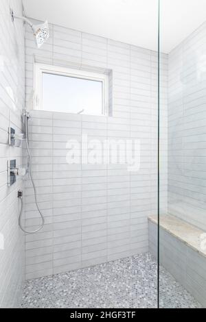 Une douche carrelée grise avec pomme de douche chromée et tuyau, une banquette et de petits carreaux circulaires sur le sol. Banque D'Images