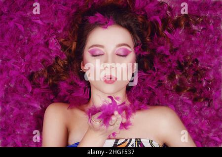 Une belle fille avec un maquillage violet professionnel envoie un baiser d'air à l'appareil photo.Le modèle est en plumes violettes.Photo de mode sur un backgrou rose Banque D'Images