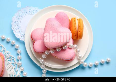 Assiette avec de savoureux macarons en forme de cœur et des perles sur fond bleu Banque D'Images