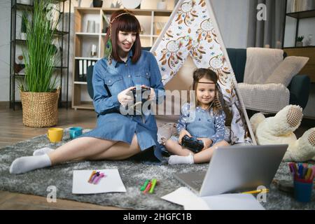 Un petit enfant heureux et une mère agréable utilisant un ordinateur portable moderne et des joysticks pour jouer à des jeux vidéo à la maison.Famille caucasienne de deux personnes assises sur le sol près du wigwam jouet. Banque D'Images