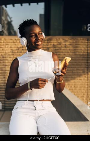 Une femme afro-américaine aime écouter de la musique avec un téléphone portable et un casque tout en étant assise à l'extérieur. Banque D'Images