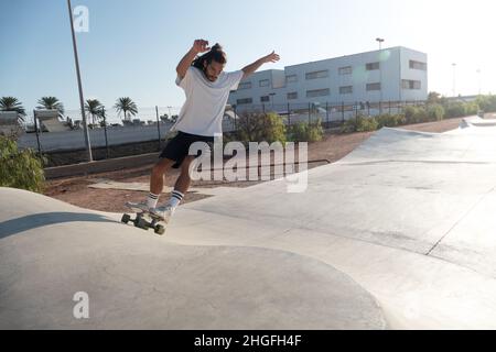 Patineuse masculine jeune adulte sur les rampes du parc de skate.Il porte des vêtements d'été et il a de longs cheveux noirs. Banque D'Images