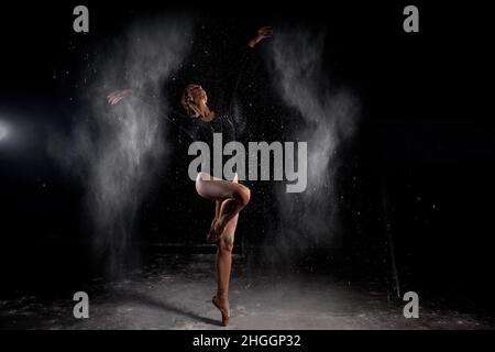 belle danseuse de ballet élancée portant un body noir, pose sensuelle parmi la farine volante qui couvre son corps, sur fond noir studio. AR Banque D'Images
