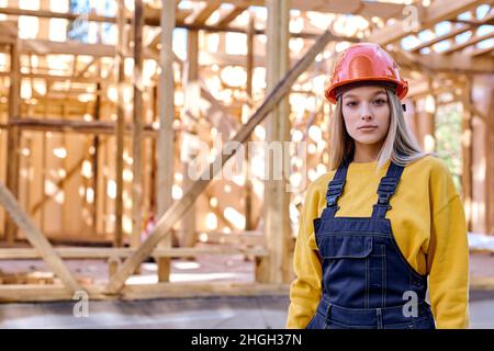 Portrait d'une jeune femme d'architecte de constructeur qualifiée en casque résistant posant contre la nouvelle construction résidentielle maison de charpente, à la journée ensoleillée dehors, habillé Banque D'Images