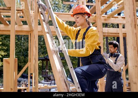 Idées de rénovation et d'extension de maisons en bois.Femelle sur échelle, monte jusqu'au toit.Travailleur Caucasien Builder en uniforme de travail et sécurité ha Banque D'Images