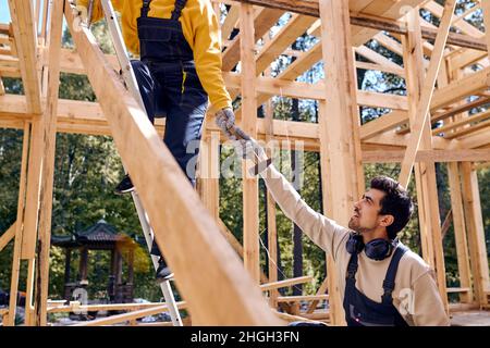 l'homme barbu en uniforme aide le constructeur féminin debout en hauteur sur l'échelle, l'homme barbu réactif donne le marteau, travaillant en équipe. amical et Banque D'Images
