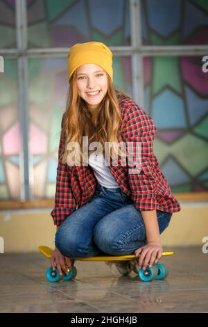 Portrait vertical de style de vie en plein air d'une jeune fille adolescente souriante avec un skateboard jaune portant un chapeau jaune et une chemise à carreaux rouge Banque D'Images