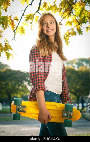 Portrait vertical de style de vie en plein air d'une jeune fille adolescente souriante avec un skateboard jaune portant une chemise à carreaux rouge Banque D'Images