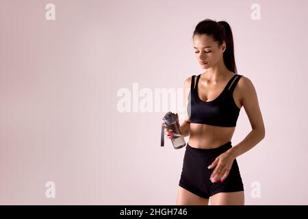 Une femme athlétique mince et soif veut boire après un entraînement actif, tient une bouteille d'eau, une femme fatiguée est habillée de vêtements de sport noirs, isolée sur une co rose Banque D'Images