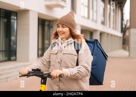 Joyeuse femme livraison de la nourriture de messagerie avec grand sac à dos thermique sur scooter électrique sur la rue de ville. Banque D'Images