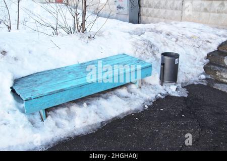Un vieux banc sale avec de la peinture écaillée, sur fond de neige sale et une poubelle. Les concepts d'un environnement urbain instable, la dévastation, la vie dysfonctionnelle. Banque D'Images