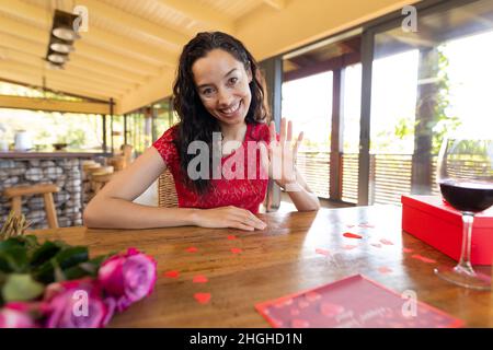 Portrait d'une jeune femme biraciale souriante avec vin et cadeau qui agite la main dans le restaurant Banque D'Images