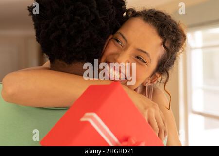 Jeune femme biraciale excitée tenant le présent de Valentin embrassant l'ami afro-américain à la maison Banque D'Images