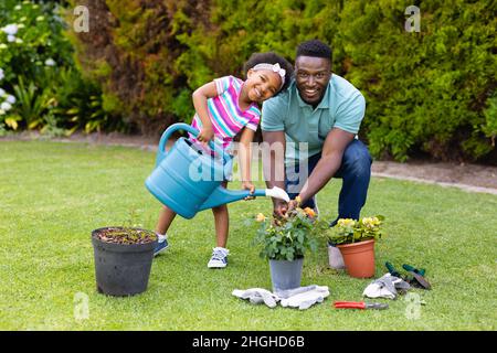 Portrait d'une fille afro-américaine souriante arrosoir des plantes par un père s'agenouillant dans le jardin Banque D'Images