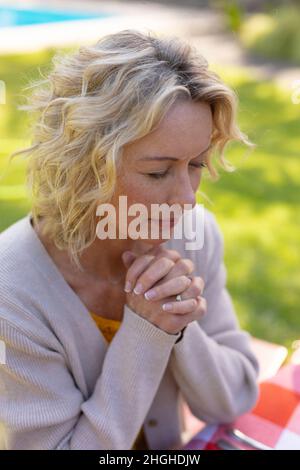 Femme blanche blonde âgée avec les yeux fermés et les mains jointes priant dans le jardin Banque D'Images