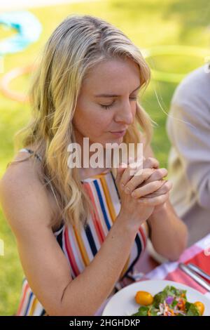 Femme blanche blonde avec les mains jointes et les yeux fermés priant à la table Banque D'Images