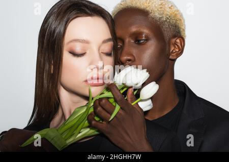 homme afro-américain blond tenant des tulipes blanches près de jolie femme avec les yeux fermés isolés sur le gris Banque D'Images