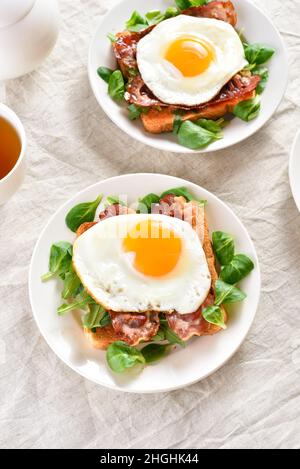 Des sandwichs ouverts avec des œufs frits, du bacon et des feuilles de légumes sur une assiette sur fond clair. Banque D'Images