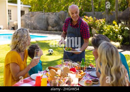 Joyeux homme de race blanche servant de la nourriture barbecue à la famille assis ensemble à table dans le jardin Banque D'Images