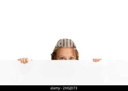 Drôle d'enfant, petite fille, élève dans des vêtements de style décontracté peeking dehors de derrière le mur isolé sur fond blanc.Concept d'enfance, émotions Banque D'Images