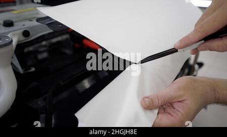 Le gabarit coupe le film sur la machine à l'aide d'un couteau.Rétromontage de la voiture avec un film de protection blanc, le maître effectue une découpe dans le revêtement avec un Banque D'Images