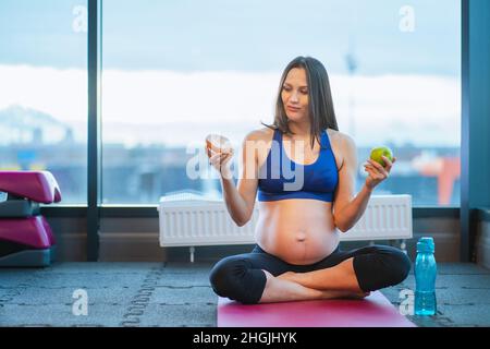 femme sportive enceinte faire le choix entre le donut et la pomme assis sur le tapis de yoga Banque D'Images