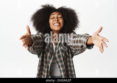 Heureuse belle femme africaine atteignant, étirant les bras à la caméra, veut se câliner, embrasser et sourire, debout sur fond blanc Banque D'Images