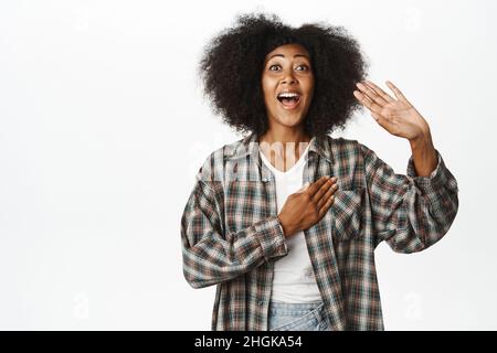 Une jeune femme afro-américaine excitée s'appelle elle-même, levant la main et agitant, en disant bonjour, salut gestuel, salutation amicale d'une amie, debout sur blanc Banque D'Images