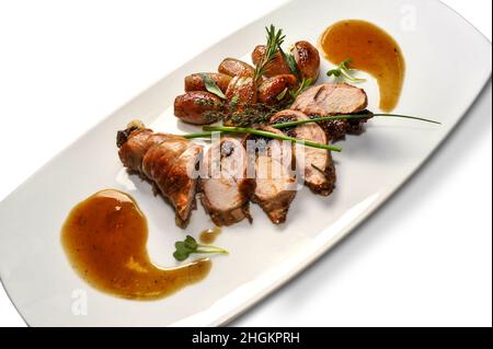 Roulé de lapin cuit farci avec pruneaux, tranches de pommes de terre et herbes aromatiques sur plaque blanche isolée sur blanc Banque D'Images