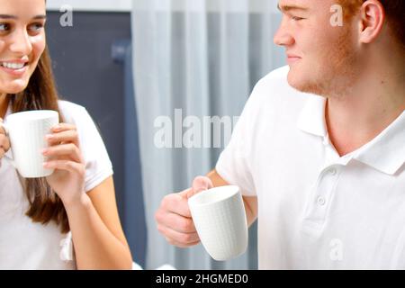 Jeune amoureux heureux couple en blanc tenant des tasses dans leurs mains le matin prenant le petit déjeuner, passant le temps ensemble. Banque D'Images